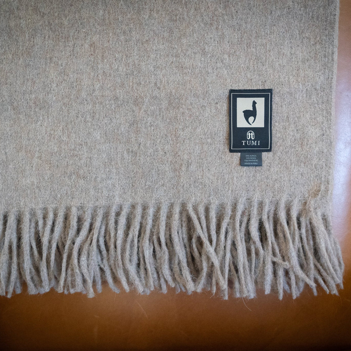 Tan alpaca wool blend throw with long fringe draped over the back of a tan leather armchair