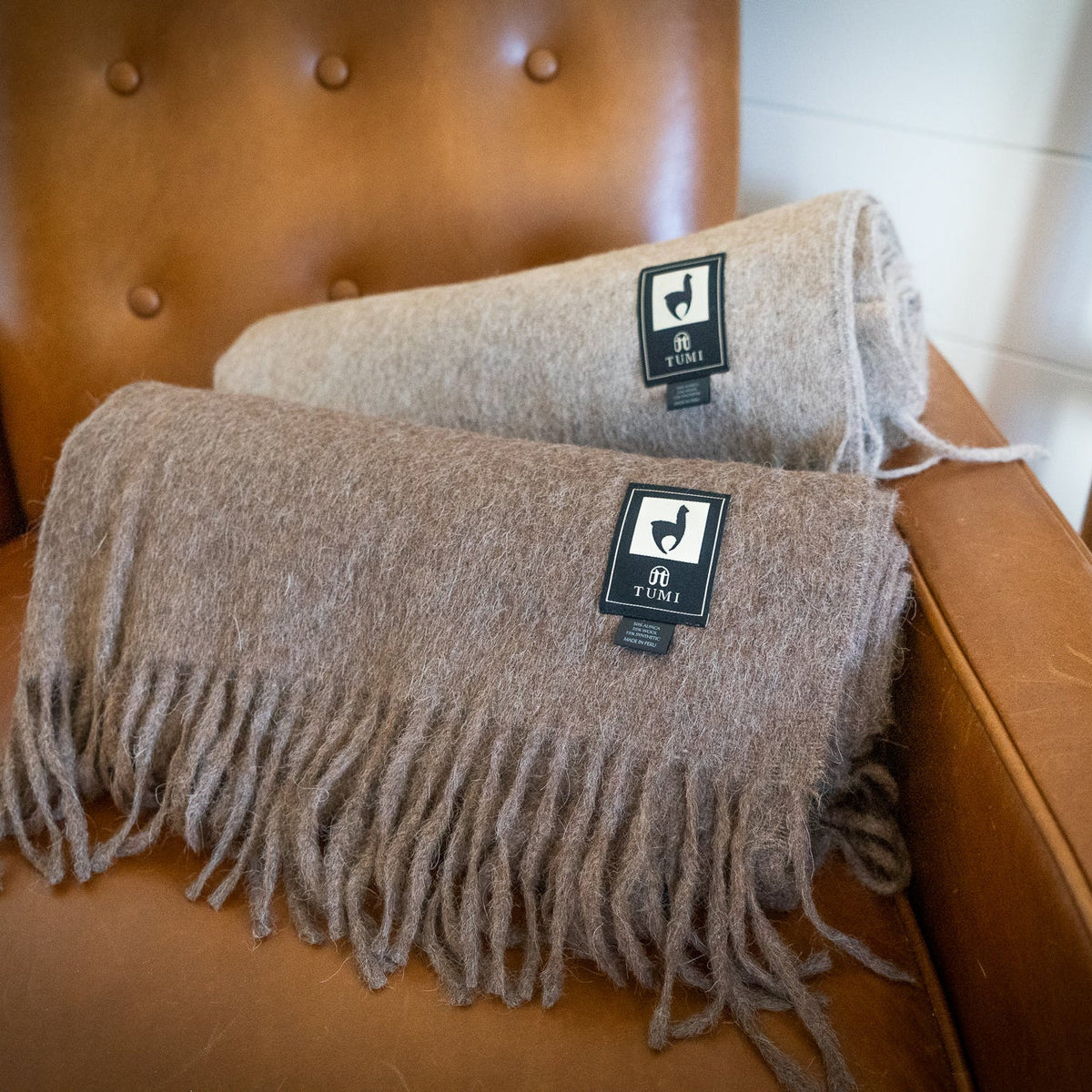 Two rolled alpaca wool blend throws in brown and tan resting on a tufted tan leather armchair