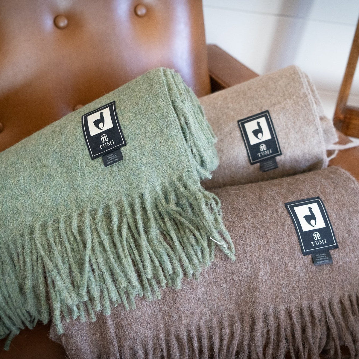 Rolled alpaca wool blend throws in green, tan and brown stacked on a tufted tan leather chair