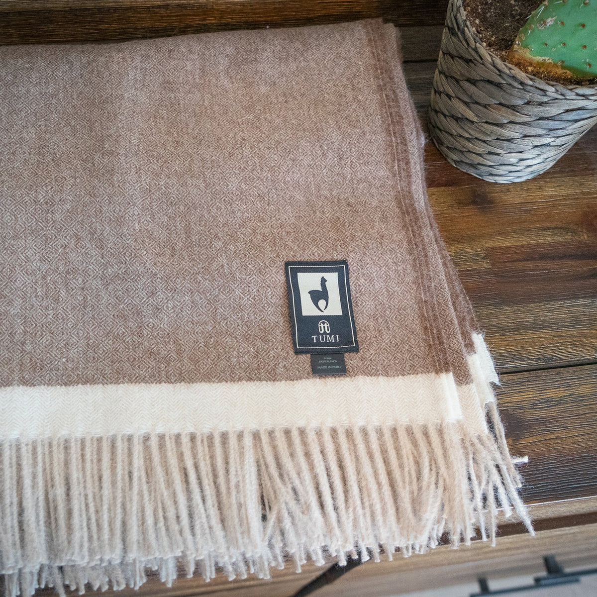 Brown baby alpaca herringbone throw folded flat on a dark wood table beside a woven basket planter