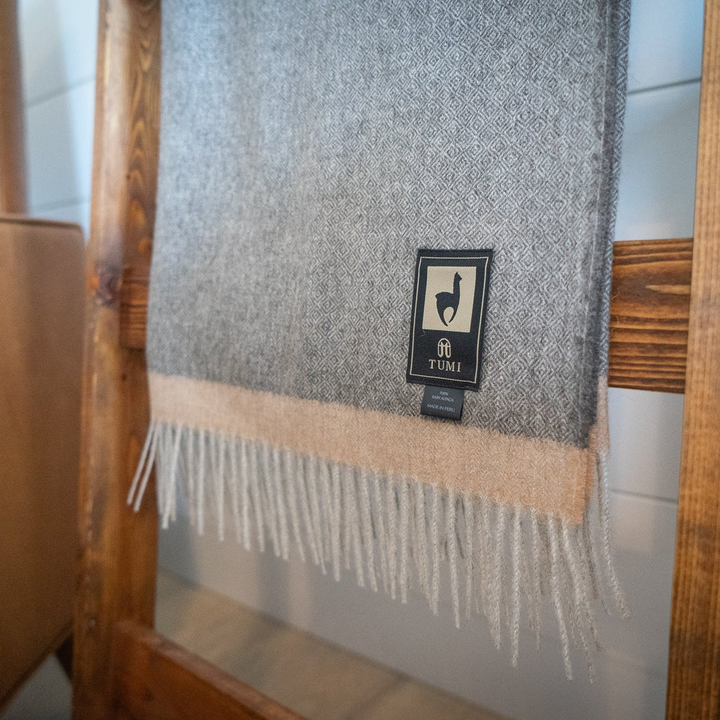 Fringed end of gray baby alpaca herringbone throw with soft tan band hanging on a wooden ladder
