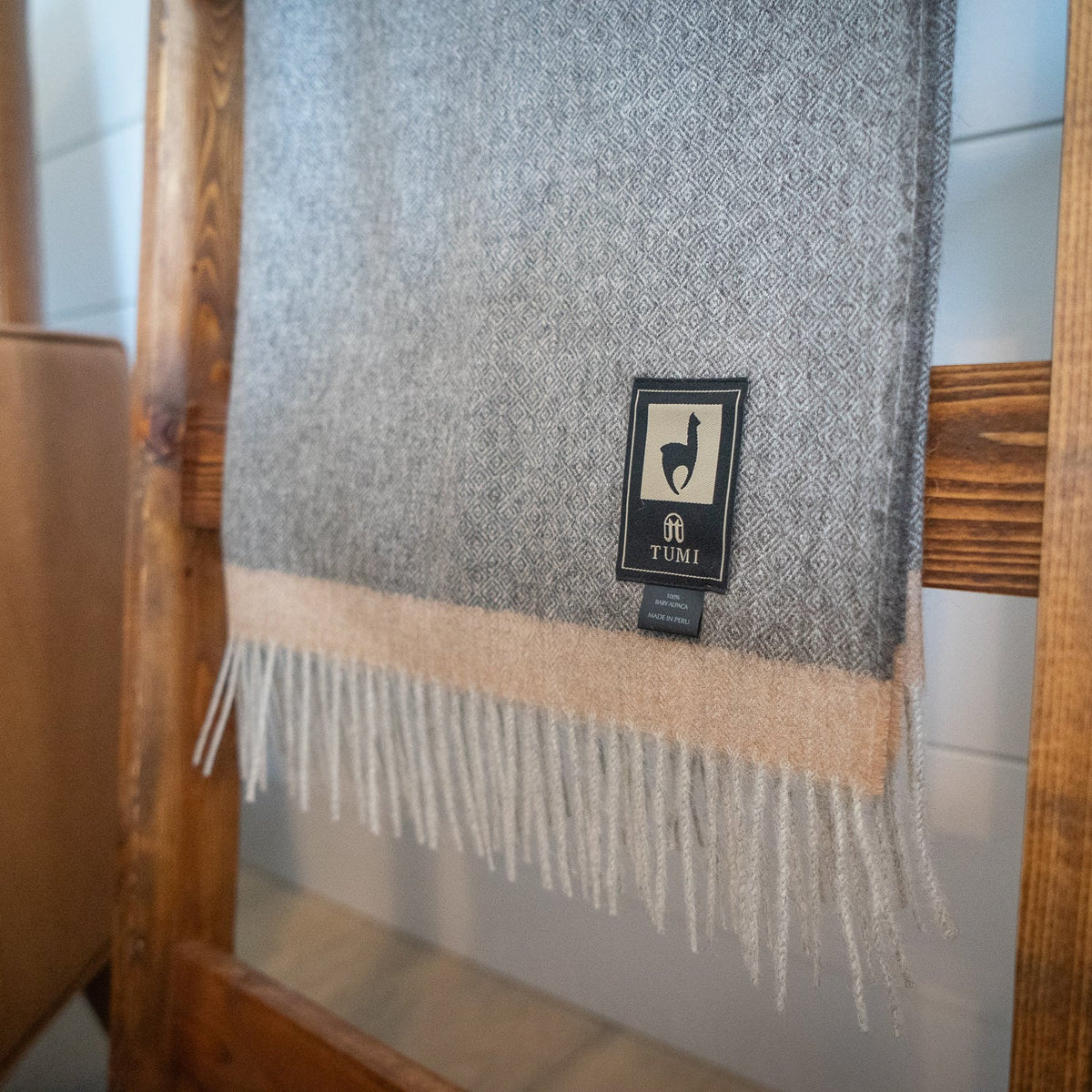 Fringed end of gray baby alpaca herringbone throw with soft tan band hanging on a wooden ladder