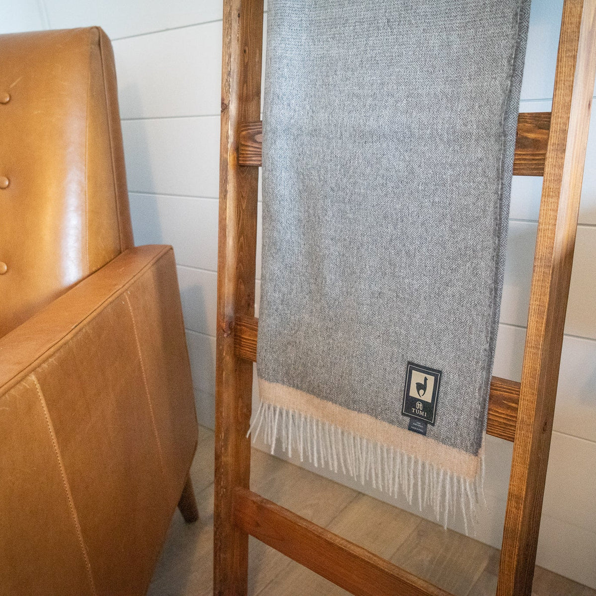 Lower half of gray baby alpaca herringbone throw with fringe hanging on a ladder near a leather chair