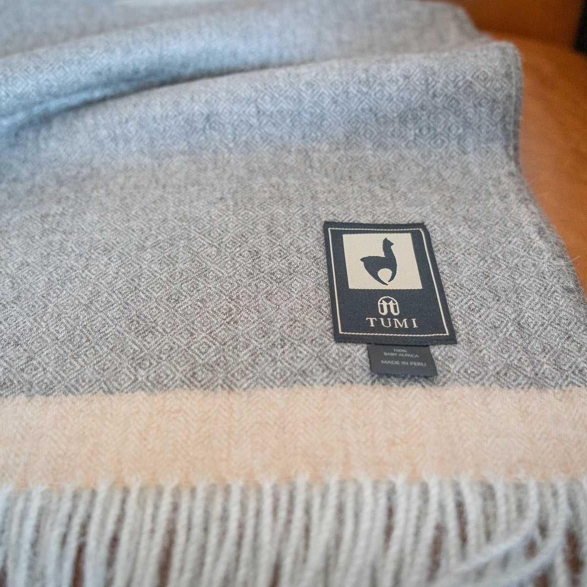 Gray baby alpaca throw with tan band and pale fringe resting on a tan leather chair, close view