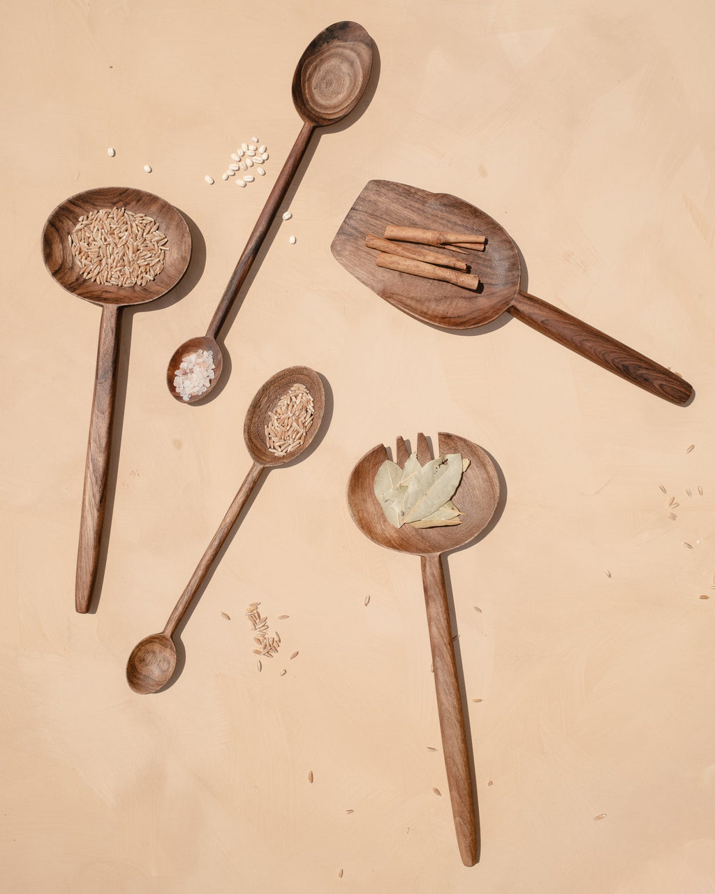 Walnut spoons, scoops and a slotted spatula holding grains, cinnamon and bay leaves on a sand-colored surface