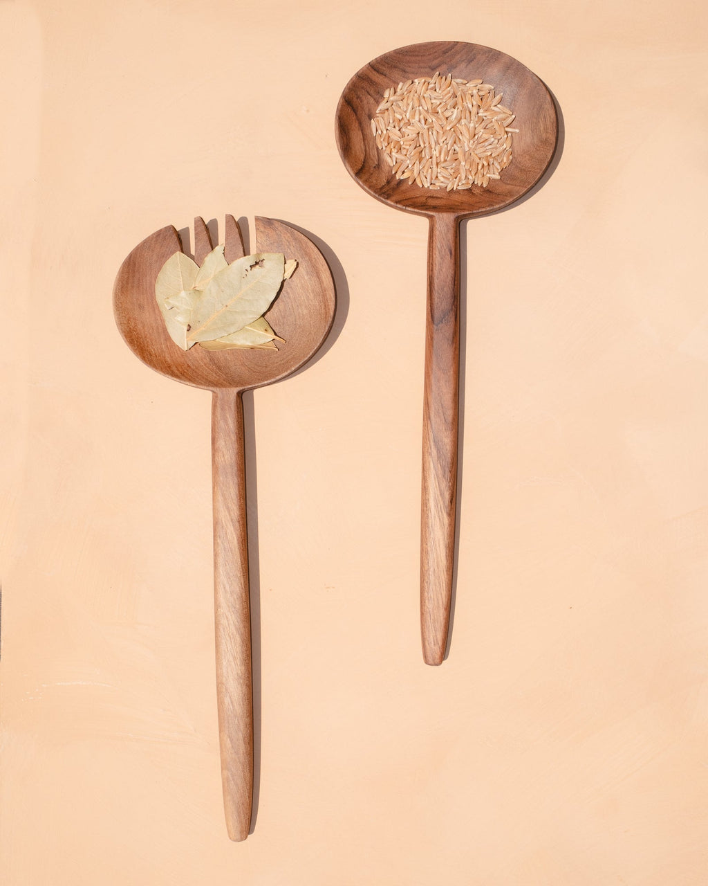 Carved walnut salad servers resting on a pale peach background, one holding a few salad leaves