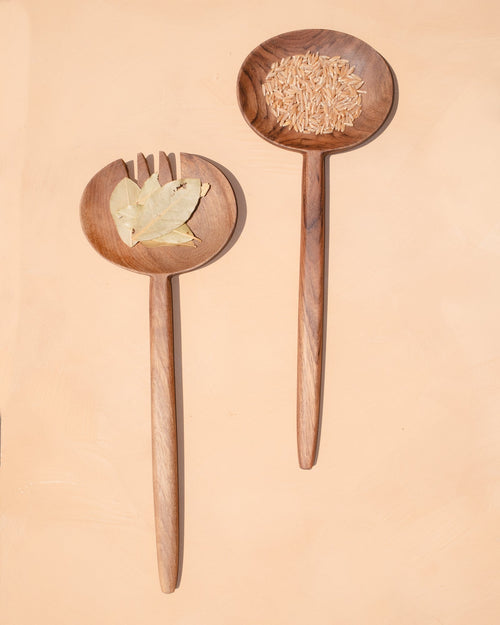 Carved walnut salad servers resting on a pale peach background, one holding a few salad leaves