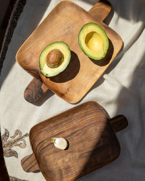 Two walnut trays from above on a white cloth, one holding a halved avocado, the other a single garlic clove