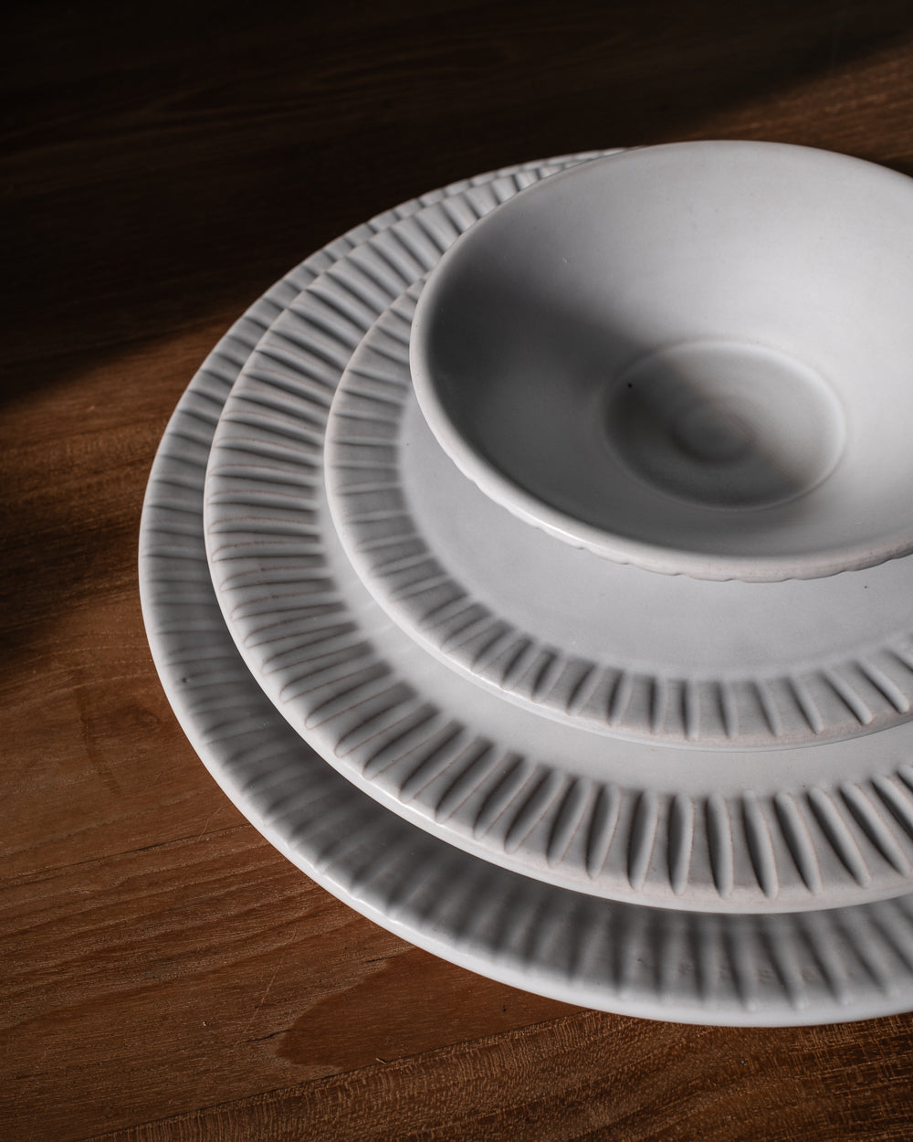 Stack of matte white carved-rim plates and a bowl in raking light, showing the hand-cut line detail