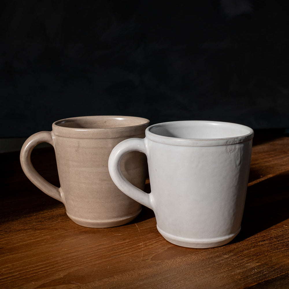 Handmade matte white ceramic mug beside a matching beige mug on a wooden table with a dark background