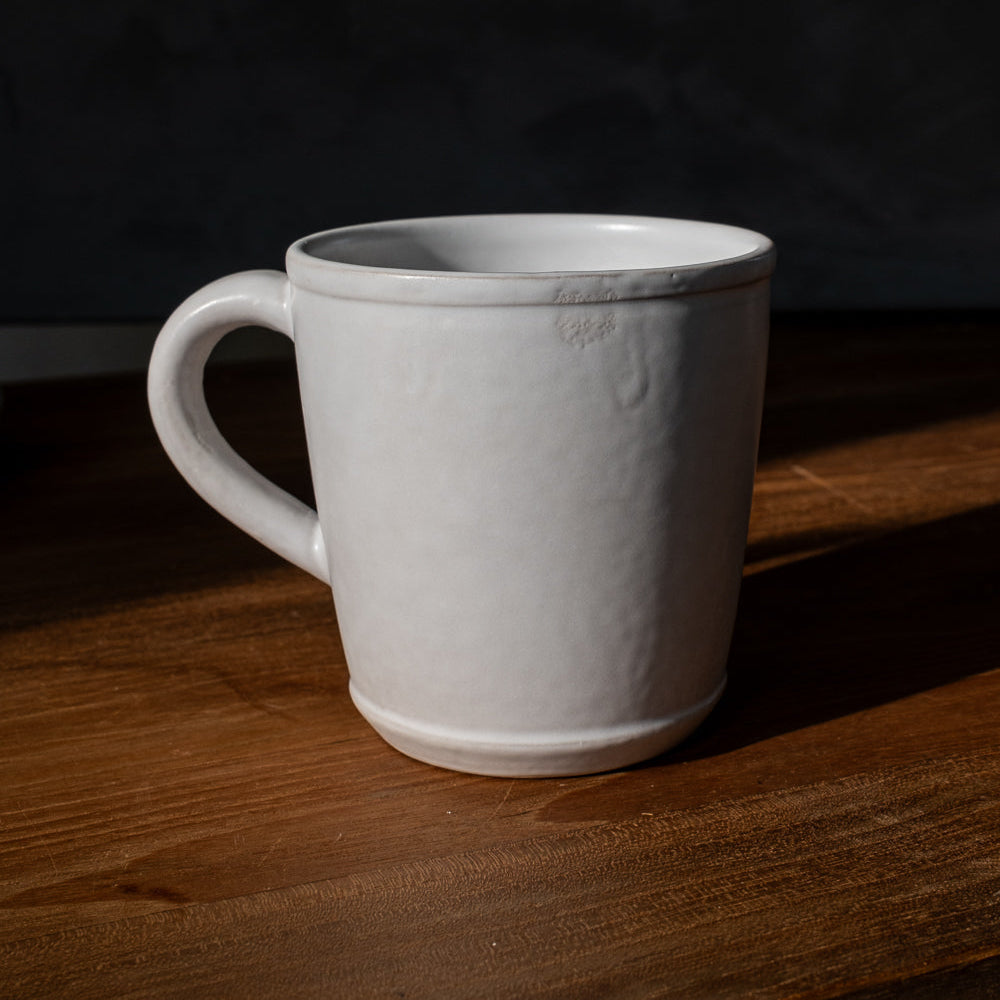 Handmade oval matte white ceramic mug on a dark wood table in a band of sunlight against a black backdrop
