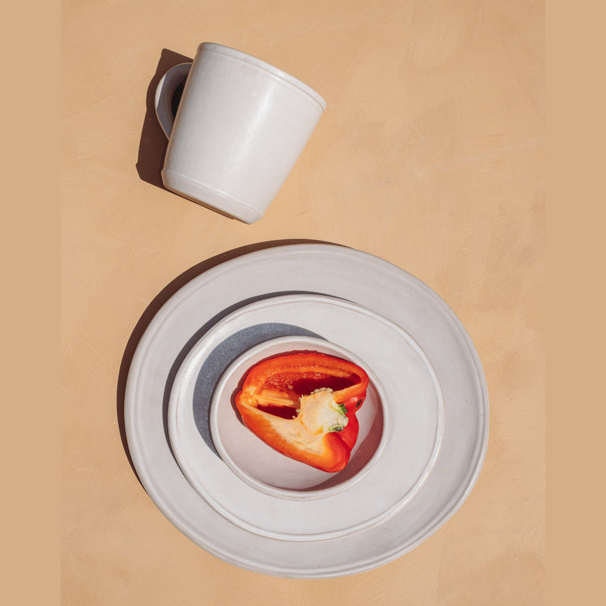 Matte white ceramic mug lying on its side above stacked oval white plates and a bowl holding half a red pepper