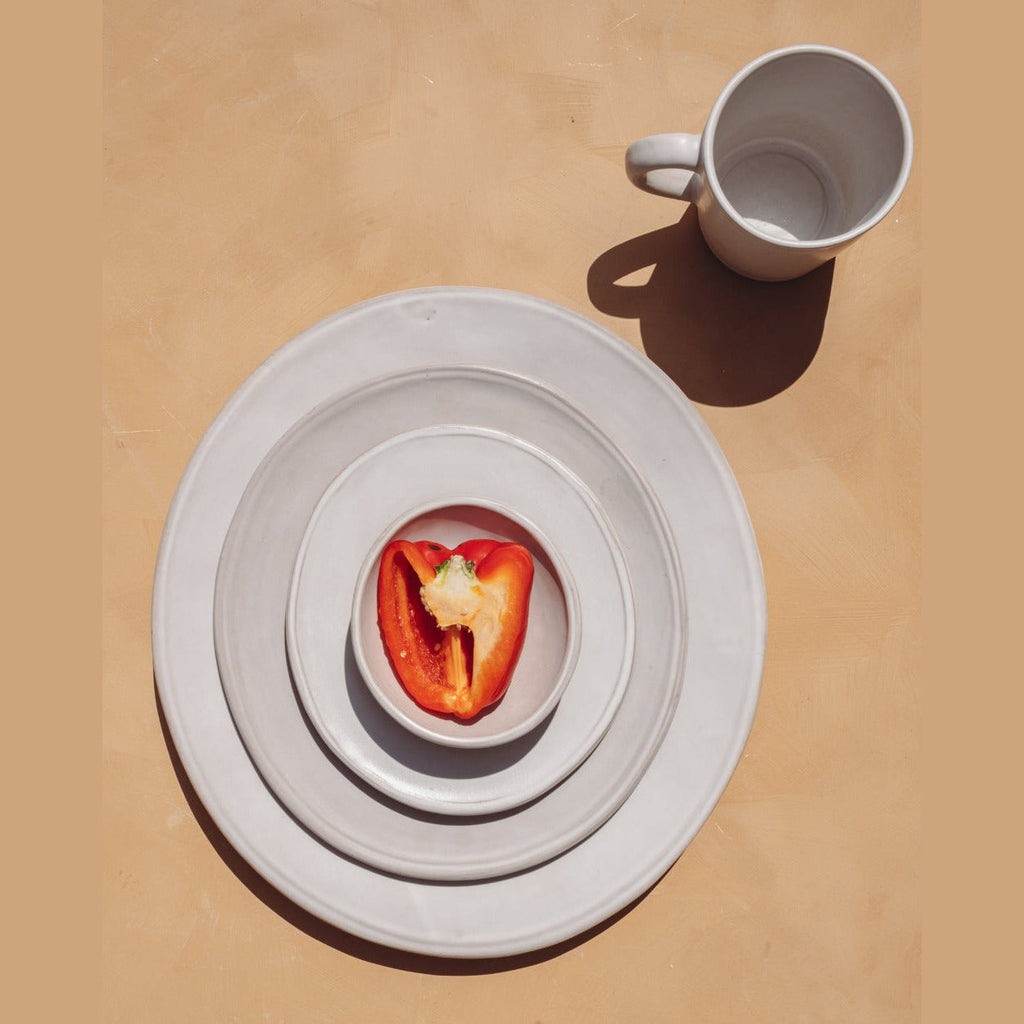 Overhead view of a matte white ceramic mug casting a shadow next to stacked oval plates with a halved red pepper