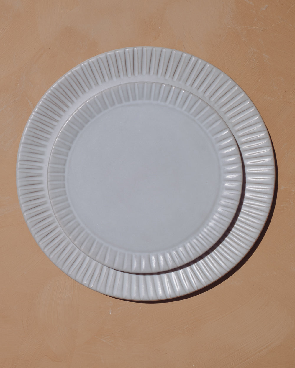 Two matte white ceramic plates with carved line rims stacked on a warm clay-colored surface from above