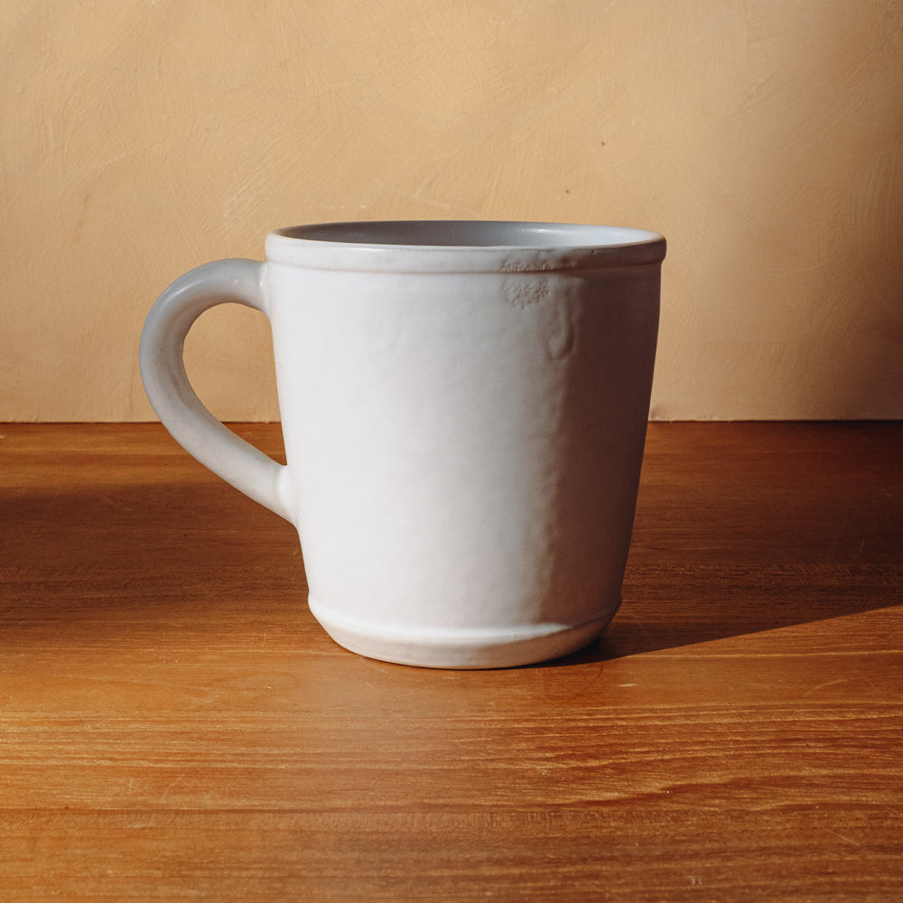 Handmade matte white ceramic mug with handle to the left on a wooden table against a warm plaster wall
