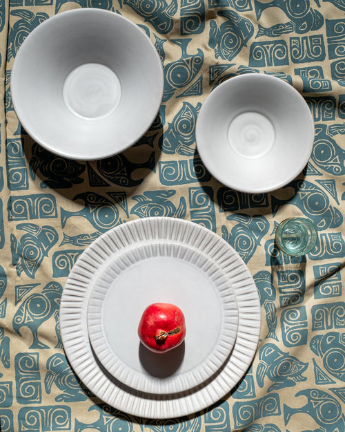 Matte white carved-rim dinner plate with matching bowls on a blue and sand printed cloth with a red apple