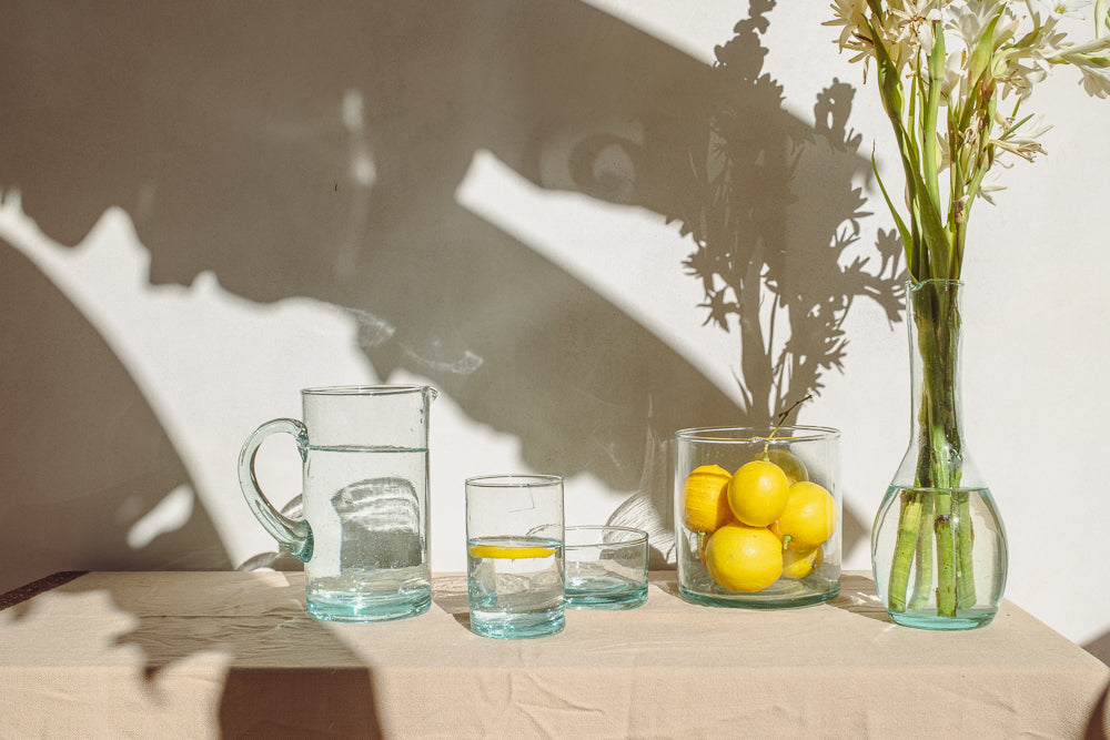 Recycled glass carafe with flowers next to a pitcher of lemons and small glasses on a sunlit table