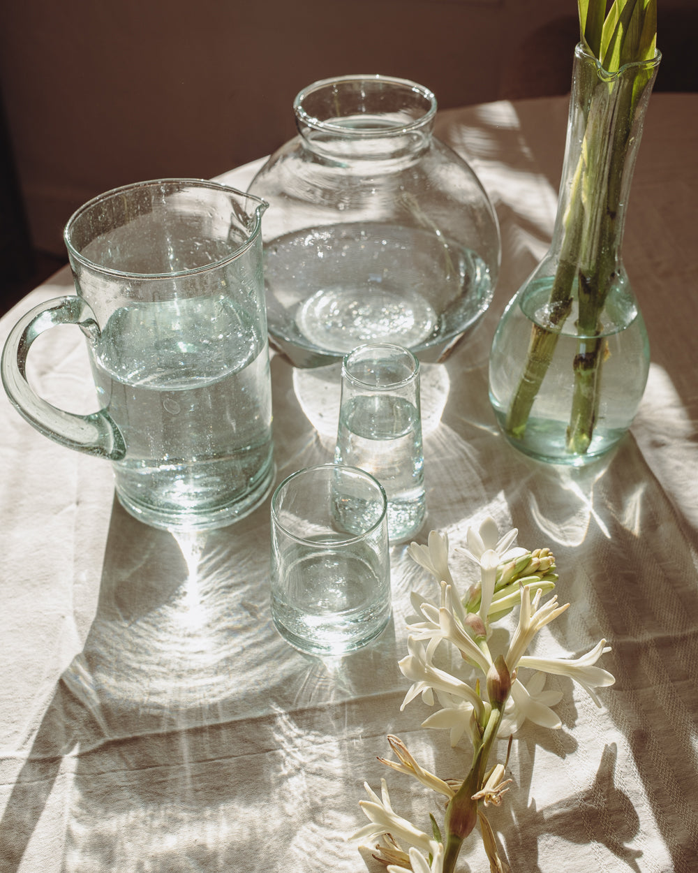 Recycled glass pitcher, carafe and tumblers on a sunny table runner with a bud vase of white flowers