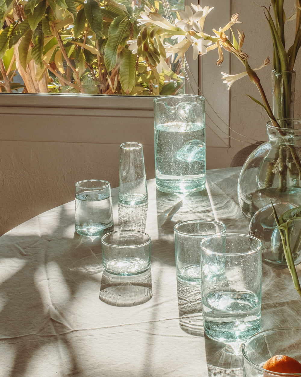 Recycled glass tumblers and a carafe on a sunlit outdoor table with dried grasses and long shadows