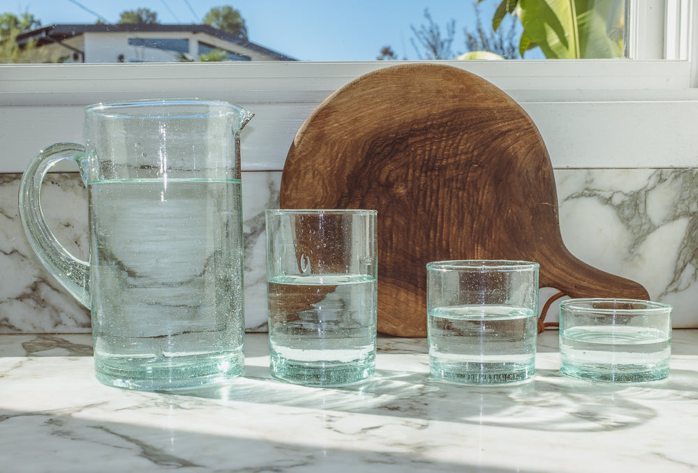 Glass pitcher and recycled glass tumblers in three sizes lined up on a marble counter by a wood board