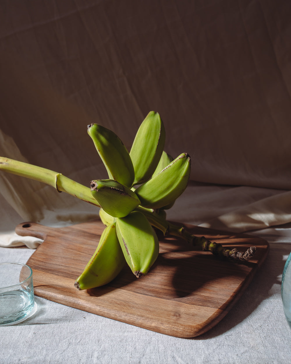 Walnut serving board on a linen-draped table holding a bunch of green bananas in late afternoon light