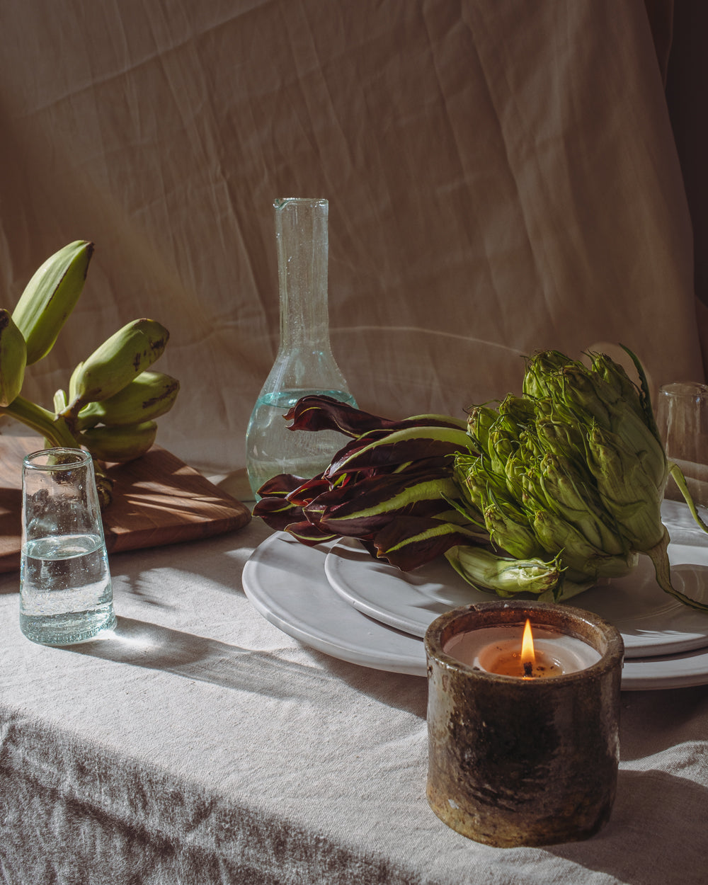 Tall green-tinted glass carafe on a linen-covered table with artichokes, plates and a lit candle