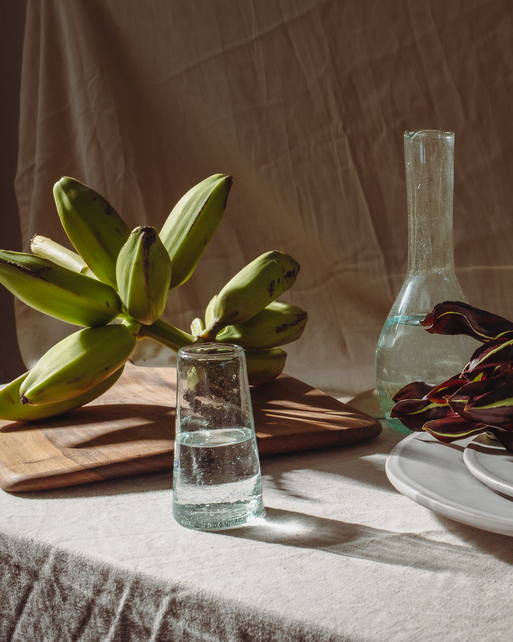 Walnut serving board on a linen table beside a glass carafe and tumbler, with green bananas in window light