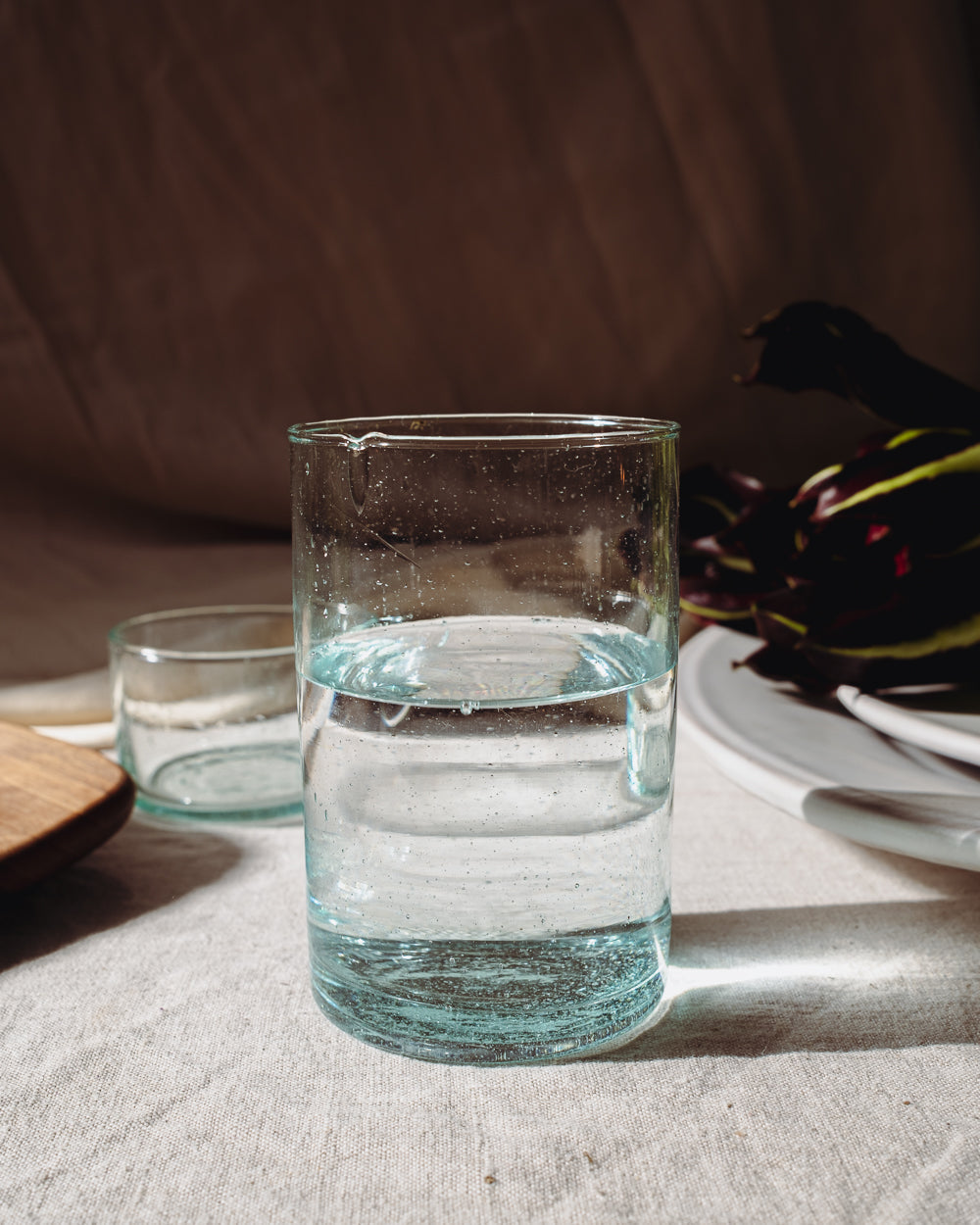 Tall recycled glass tumbler filled with water on a linen tablecloth, a small glass and dark leaves behind