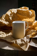 Raw stoneware pitcher with a white glazed interior standing on crumpled brown paper in warm side light