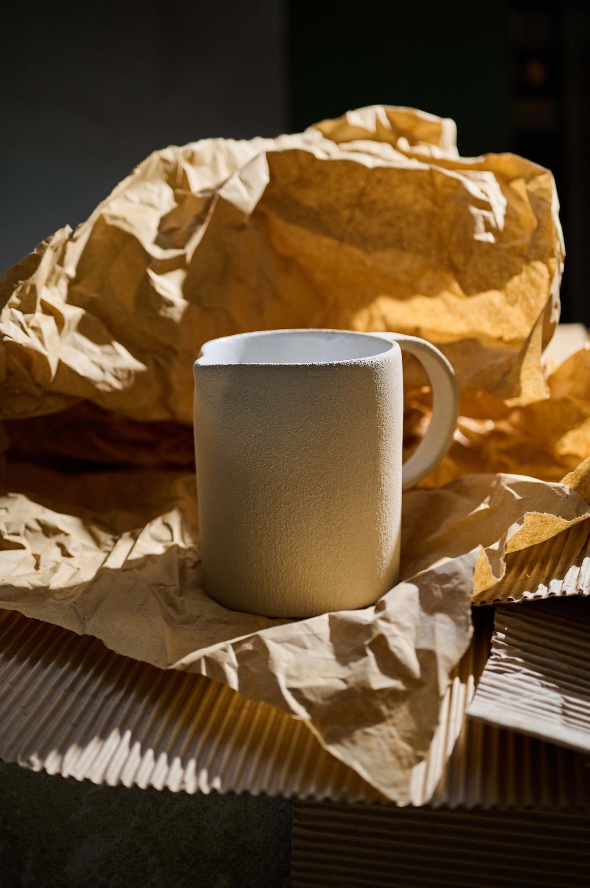 Raw stoneware pitcher with a white glazed interior standing on crumpled brown paper in warm side light