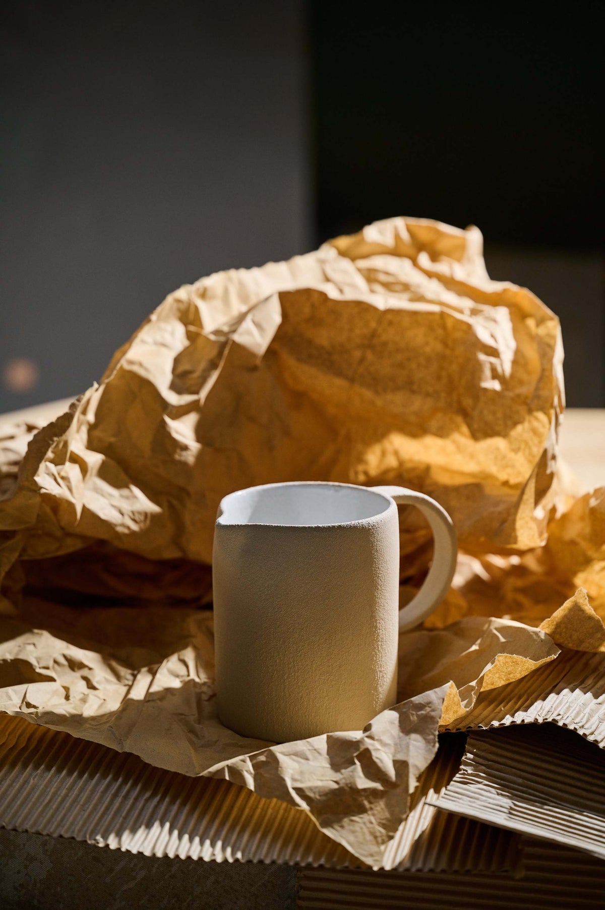 Hand-thrown stoneware pitcher in bare pale clay casting a long shadow across brown paper and corrugated card
