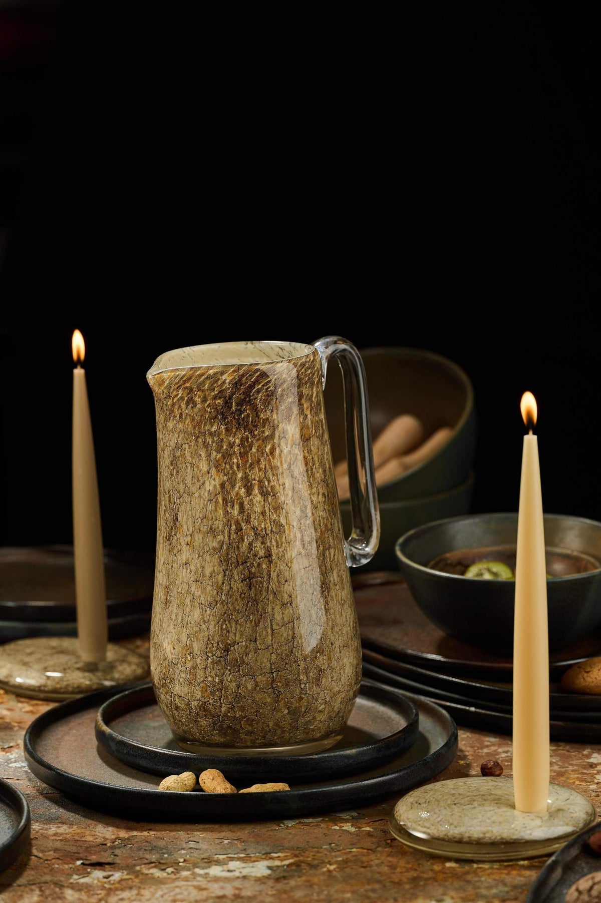 Marbled glass pitcher between two lit taper candles on a dark table set for an evening dinner