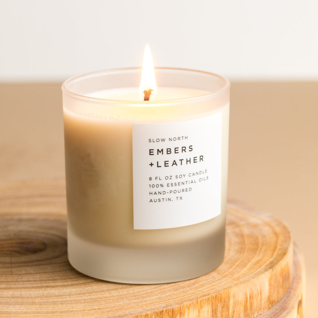Close-up of a lit frosted glass soy candle with a smoky vetiver and cedar scent on a round wood slice