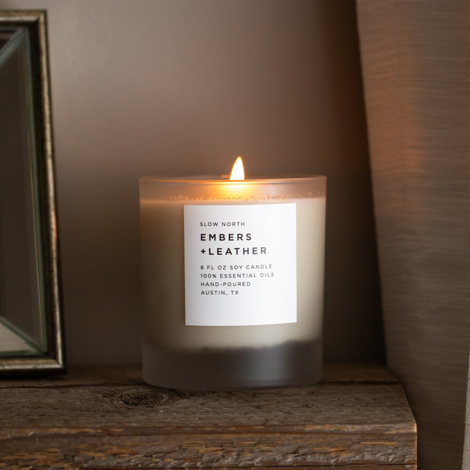 Frosted glass vetiver and cedar soy candle with a white label, lit on a wood shelf beside a dark picture frame