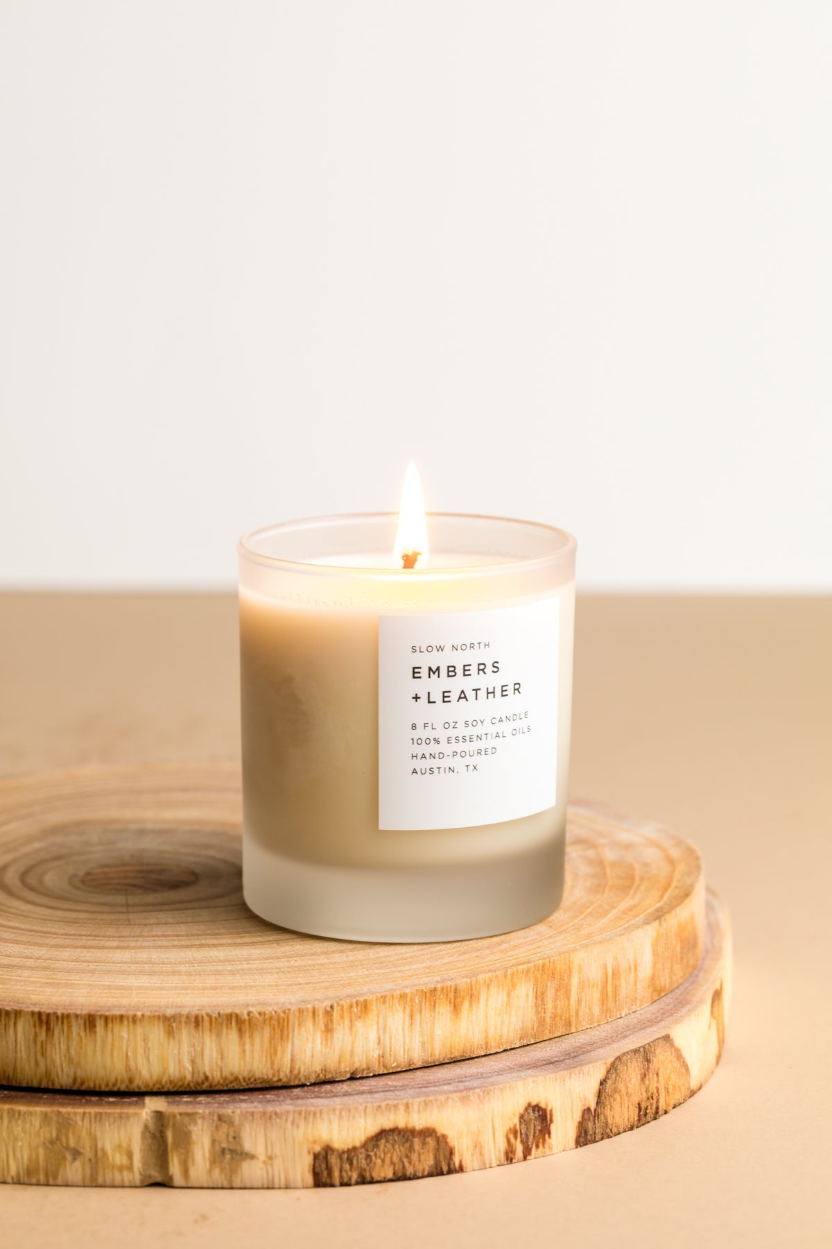Small lit frosted glass soy candle on a round wood slice against a pale cream wall in soft light