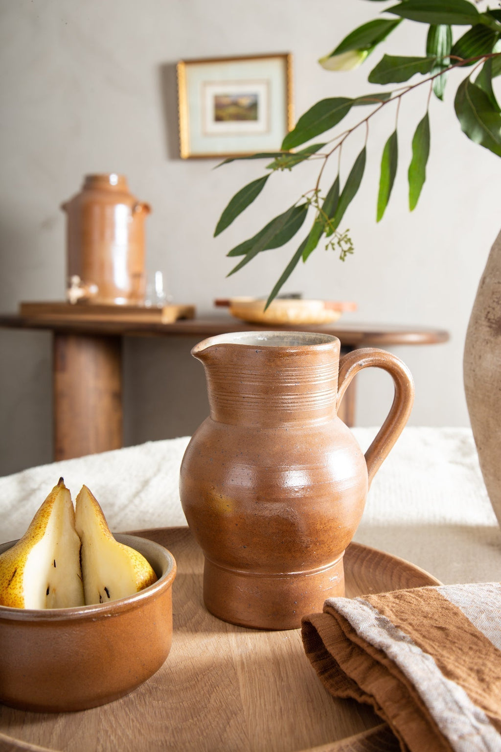 Salt-glazed French pitcher on a round wood tray beside a bowl of pears and a linen napkin in a sunny kitchen