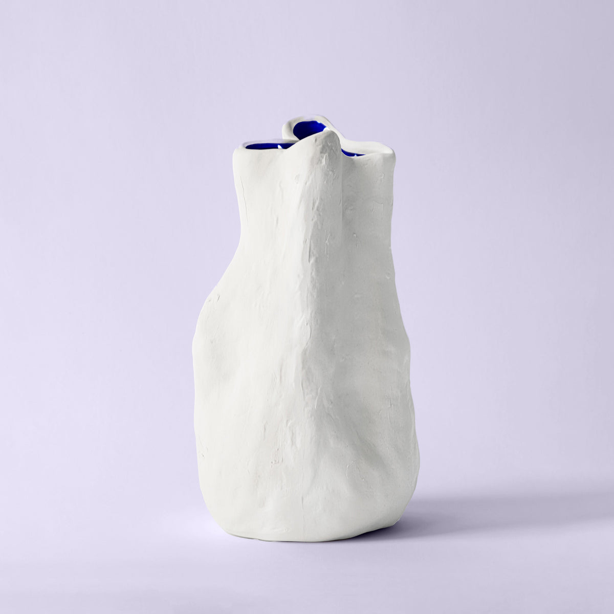 Tall white sculpted porcelain vase with leaning walls and a cobalt blue interior on a lavender backdrop