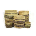 Five handwoven grass baskets in natural and black stripes, nested and stacked in a range of sizes on a white ground