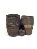Five handwoven charcoal grass baskets in graduated sizes grouped together, natural fiber flecking the dark weave