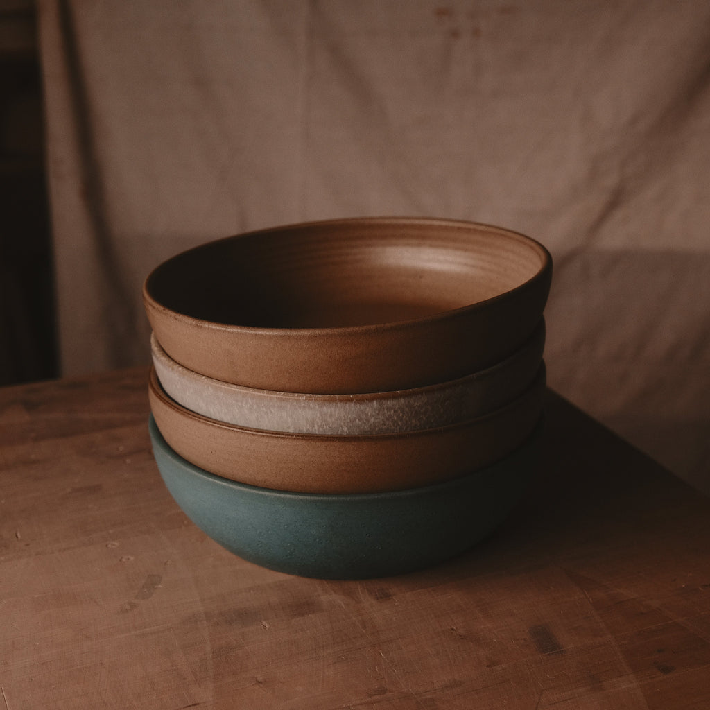 Stack of hand-thrown stoneware serving bowls in brown, oat and green glazes on a wood table against a tan wall