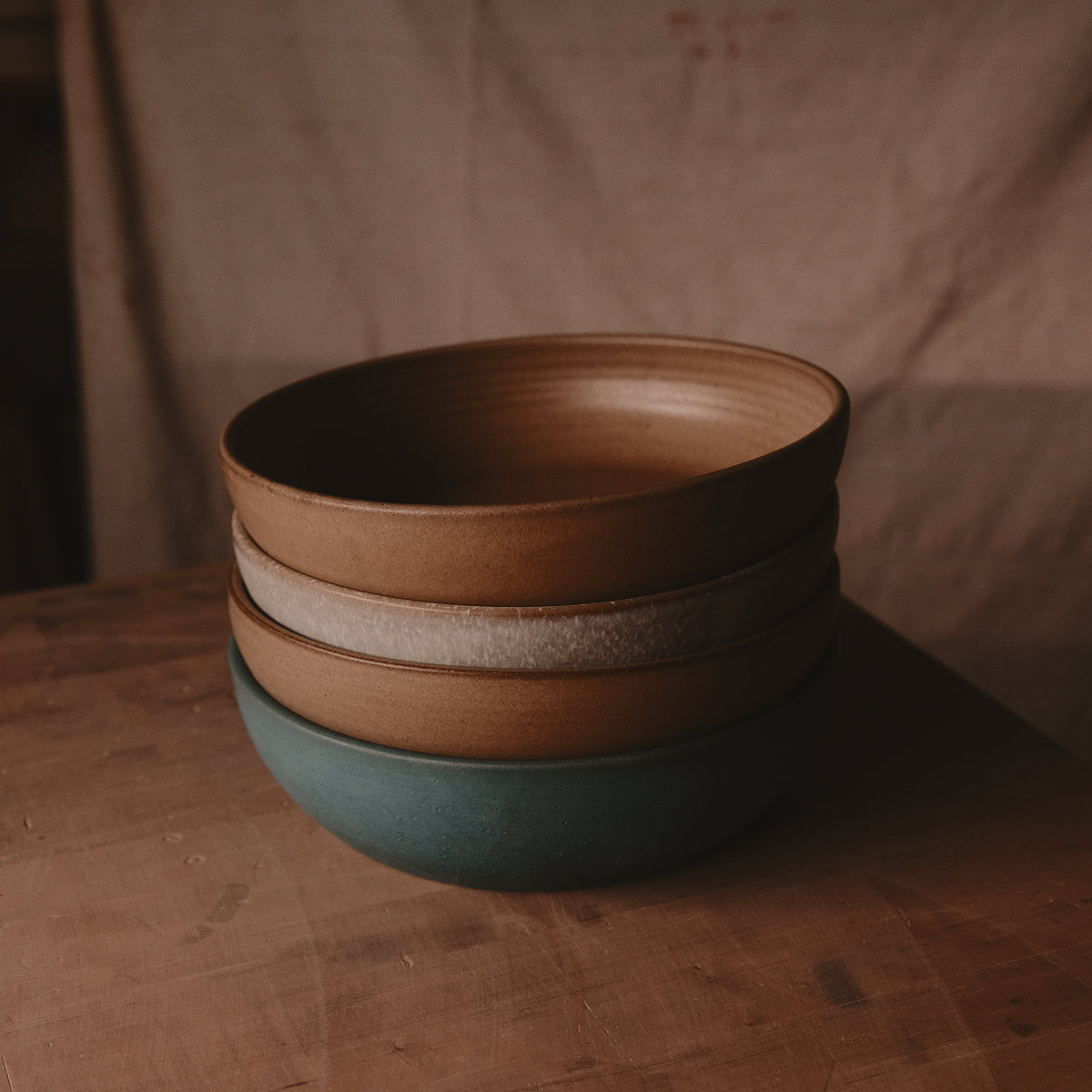 Stack of hand-thrown stoneware serving bowls in brown, oat and green glazes on a wood table against a tan wall