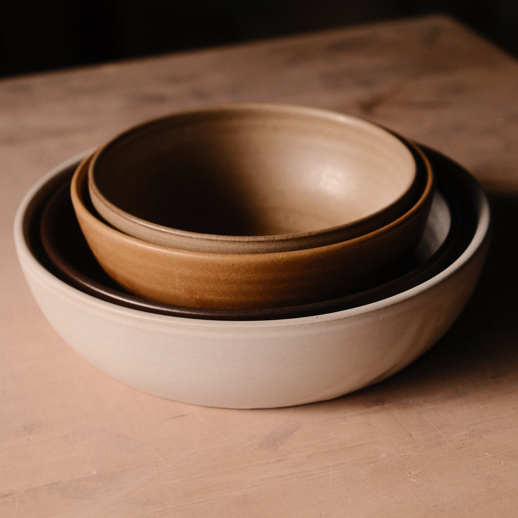 Nested hand-thrown stoneware bowls in cream, tan and brown glazes on a rosy wood surface in soft light