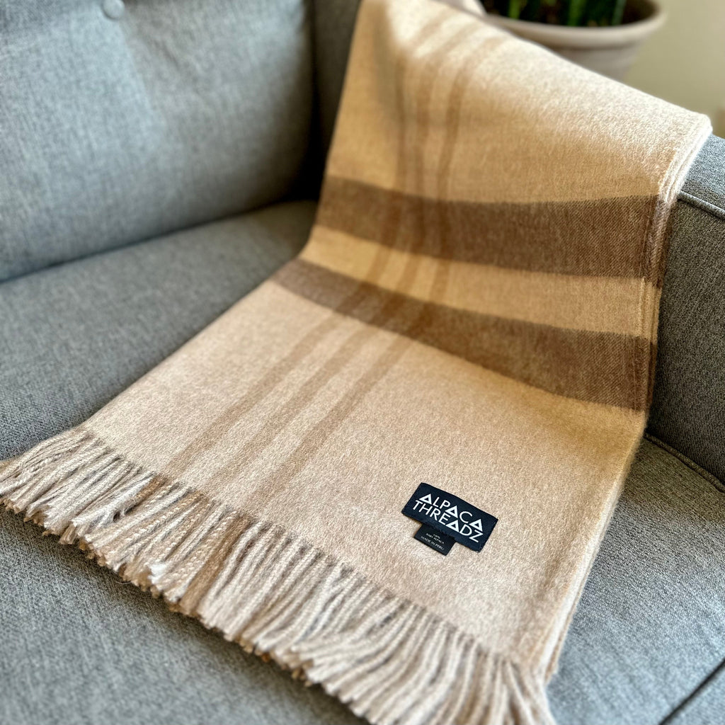 Tan and brown plaid baby alpaca throw with fringe draped over the arm of a gray tufted sofa