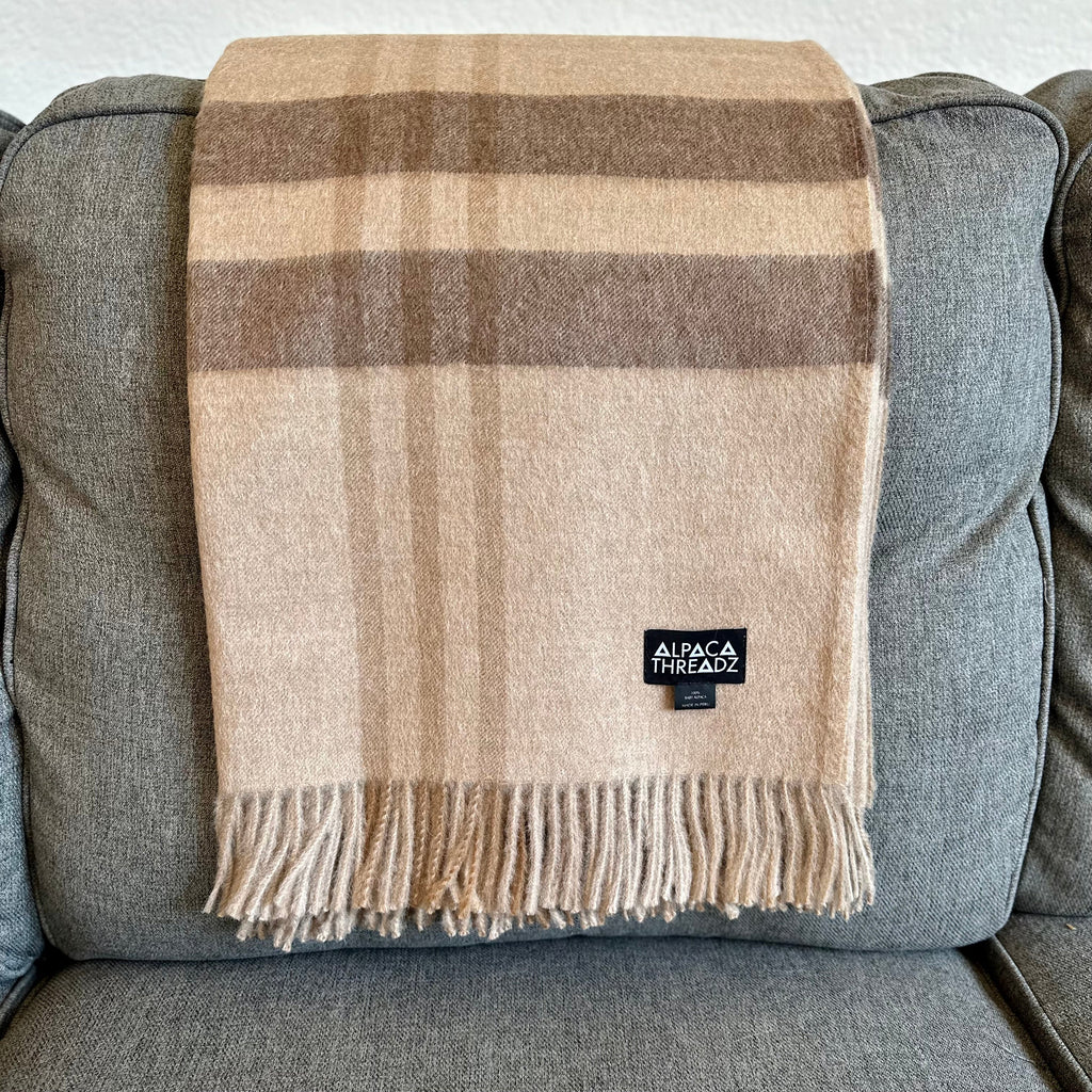 Tan and brown plaid baby alpaca throw hanging flat over the back of a gray sofa, fringe at the hem