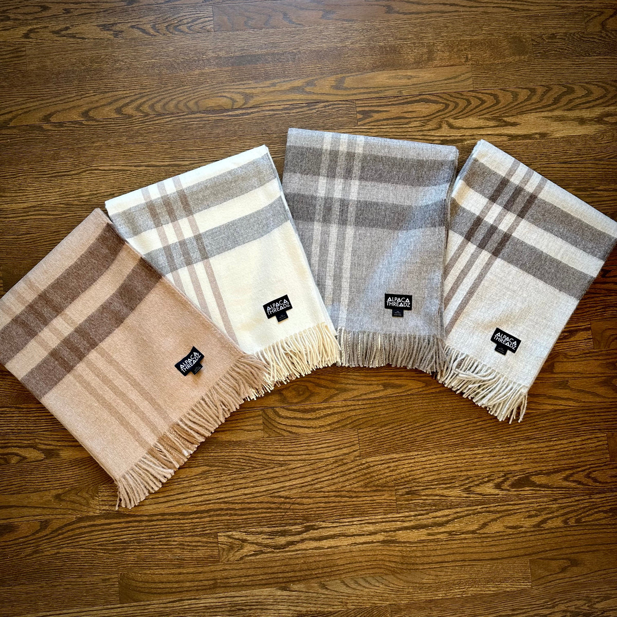 Four baby alpaca plaid throws in brown, cream and gray tones fanned out on a warm wood floor