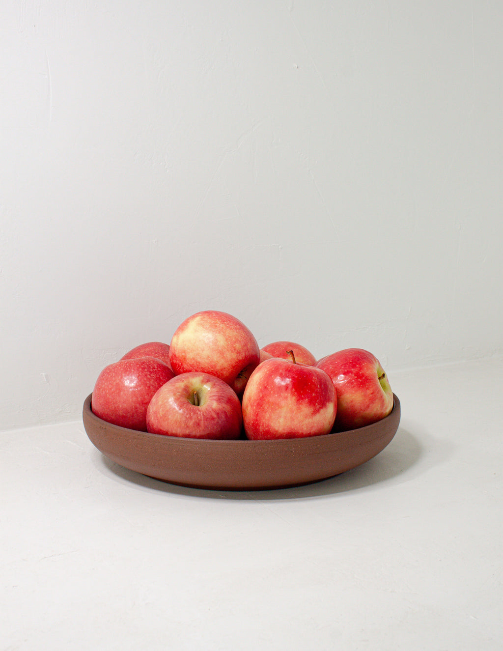 Ceramic coupe serving bowl with a bare clay base filled with red apples on a pale gray surface