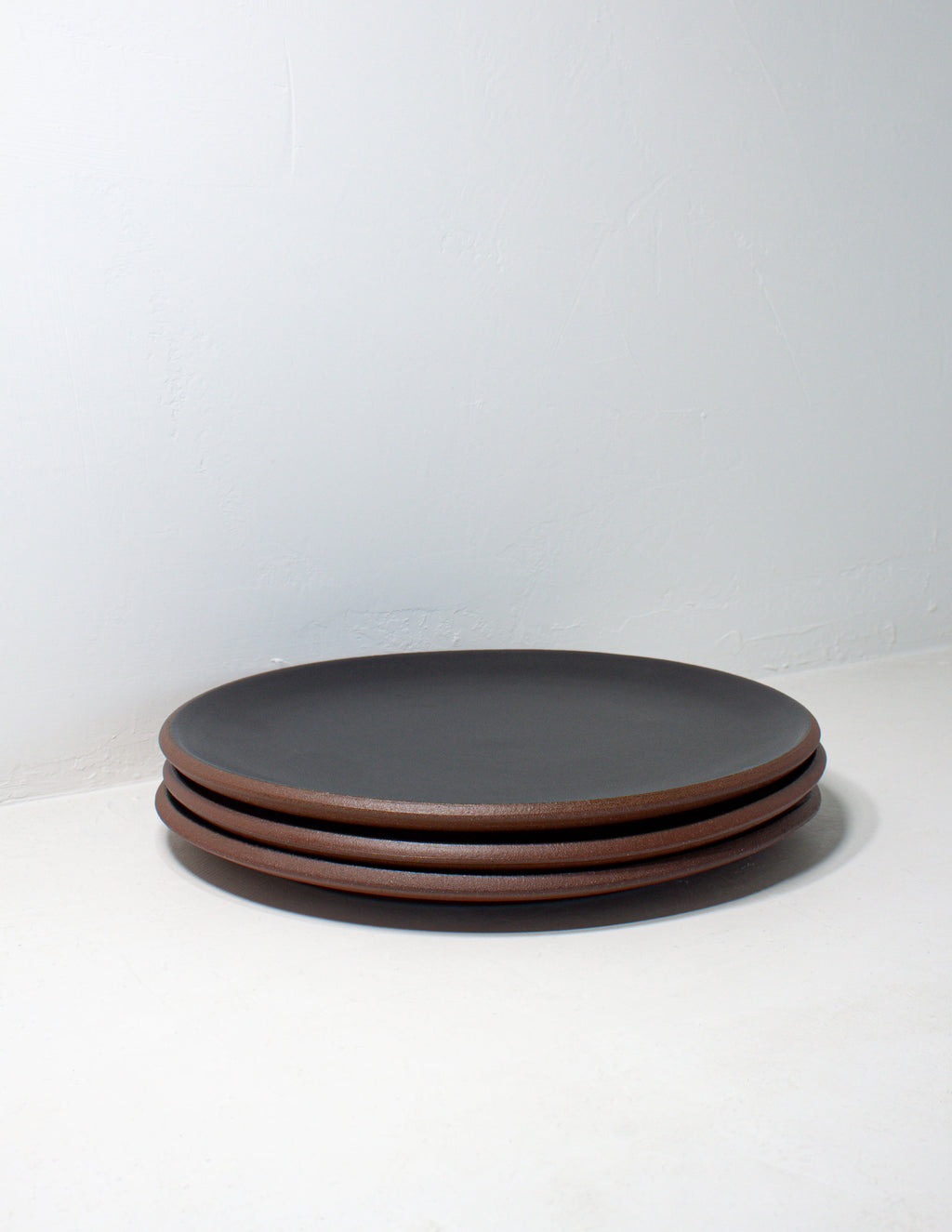 Stack of three hand-thrown stoneware coupe dinner plates in matte black glaze with bare red clay edges