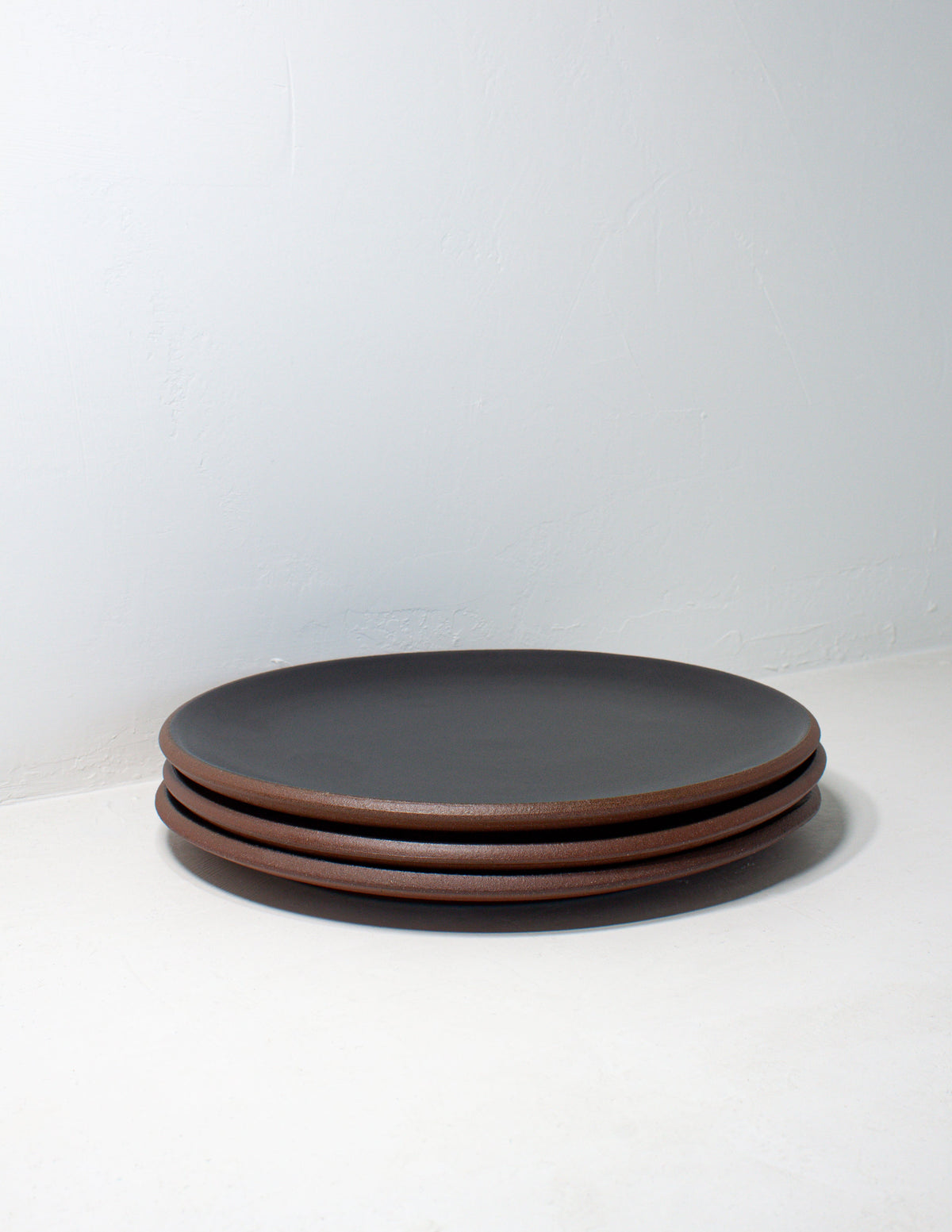 Stack of three hand-thrown stoneware coupe dinner plates in matte black glaze with bare red clay edges