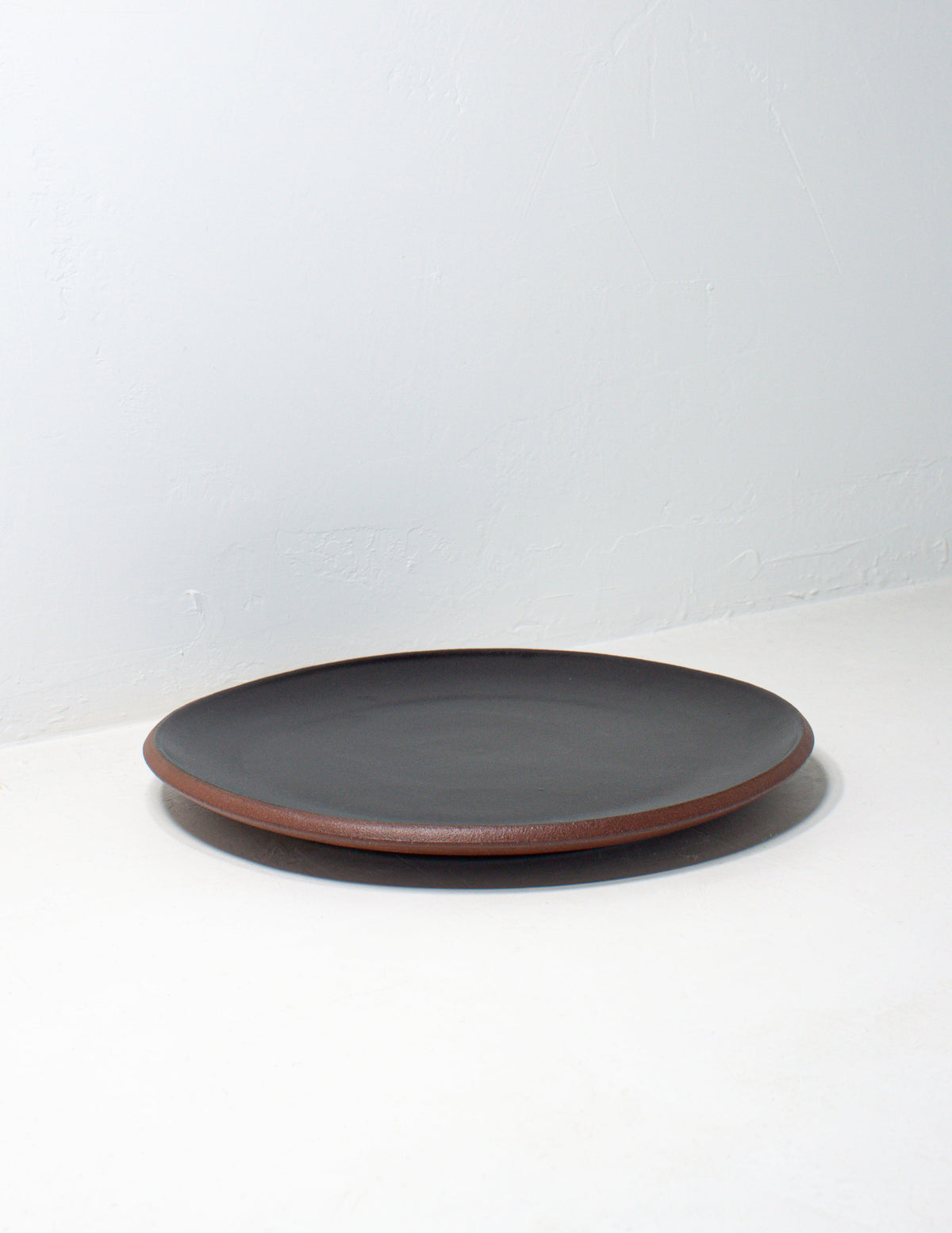 Single hand-thrown stoneware coupe dinner plate in matte black glaze lying flat, showing the red clay rim