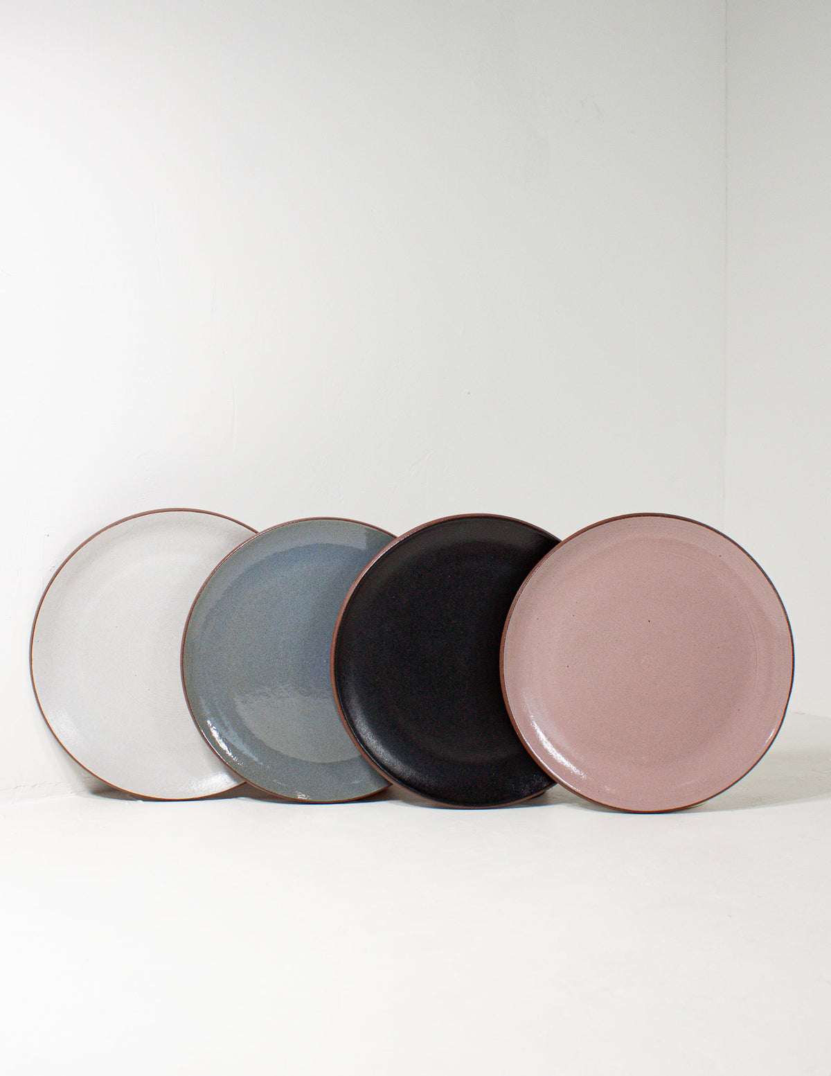 Four hand-thrown stoneware coupe salad plates in white, sky blue, black and pink glaze lined up against a white wall